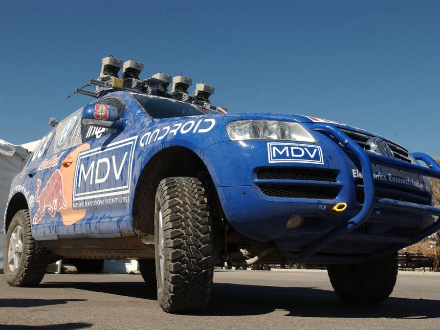 A low view of a blue car with LIDAR on top and logos on its body.