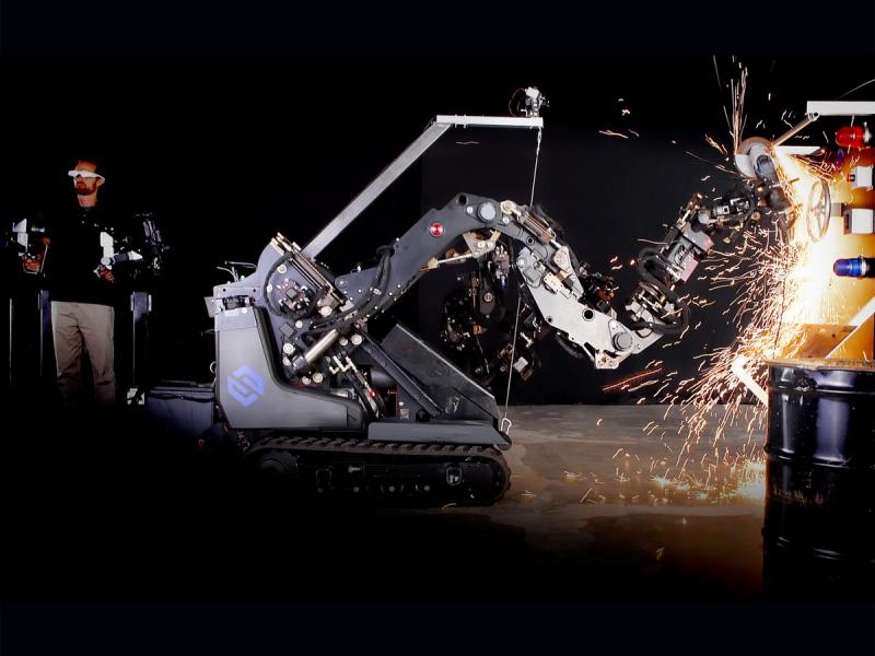 A human operator on the left wears googles and an exoskeleton system that allows him to move a large industrial robot's 7 foot long arm, which is attached to a wheeled mobile tank-like base. Sparks are flying off the end of the arm as the robot works, a safe distance from the human.