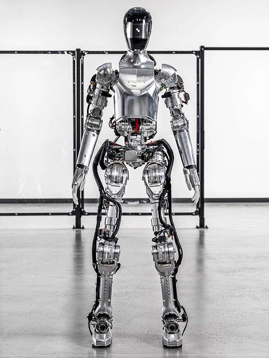 The Figure 01 robot, a shiny silver bipedal humanoid with hands and a helmeted head stands in a warehouse. 