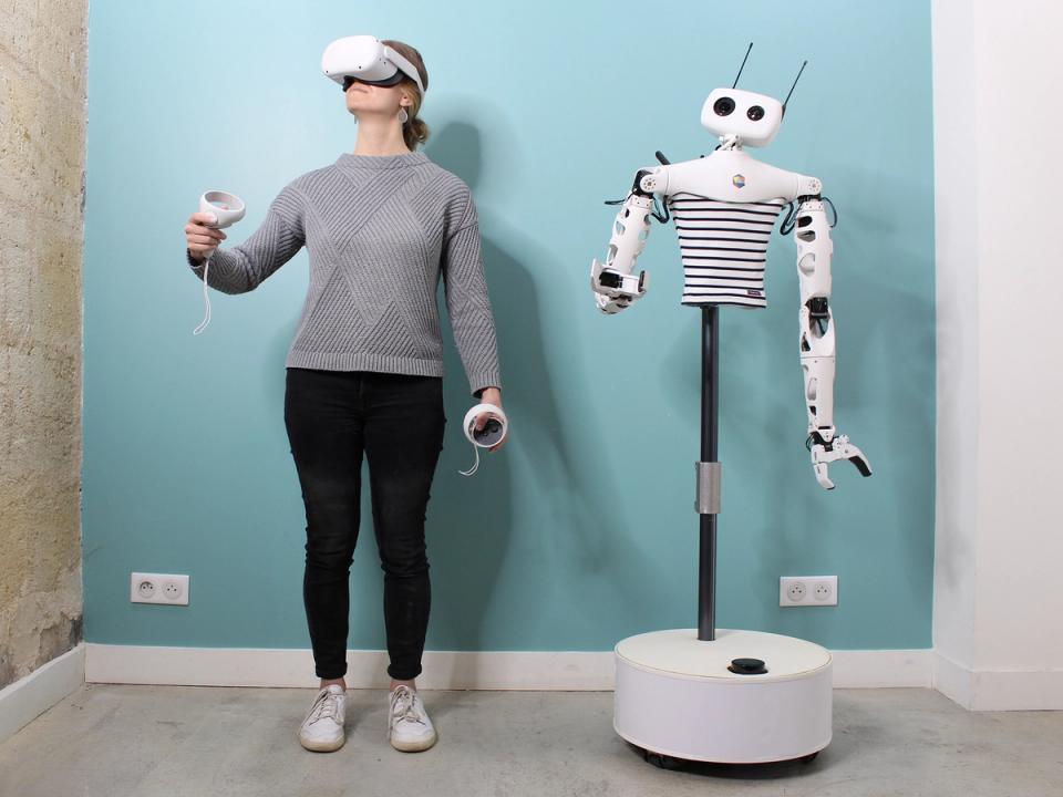 A woman wearing VR goggles and holding an implement in each hand is posed next to a humanoid torso on a pole and mobile base, which mimics her position.