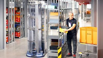 A tall mobile robot, with metal racks encased in a transparent plastic shell, bring boxes to a human worker, who picks boxes and places into a plastic case.