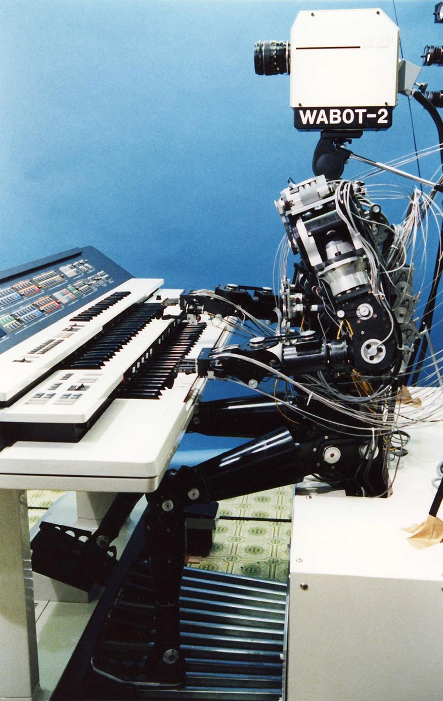 A historical robot with a complex humanoid body and a 1980's era video camera for a head sits and plays the keyboards.