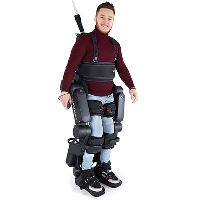 A smiling man in a dark red turtleneck and jeans in a tethered exoskeleton that includes strapping around his legs and torso.