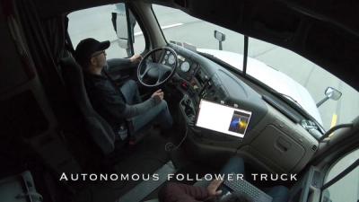 A man wearing a cap sits on the driver seat of a moving truck without touching the steering wheel while a passenger monitors a computer screen mounted on the dashboard.