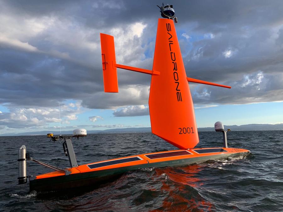 An uncrewed surface vehicle on the water. It has a horizontal orange body with multiple solar panels and a large orange sail rising vertically, and a rudder running parallel to the sea.