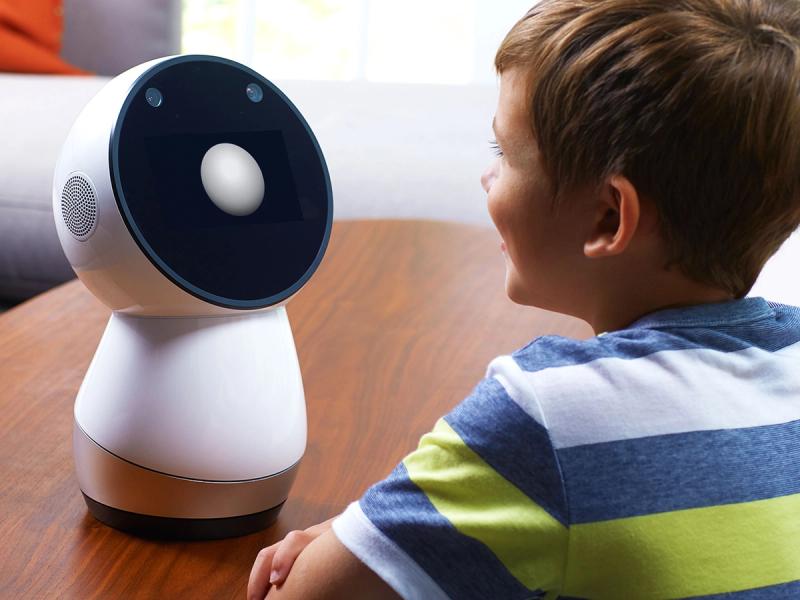 Jibo, a simple robot with a circular black display screen and bowling pin shaped white and silver base sits on a table and interacts with a young boy.