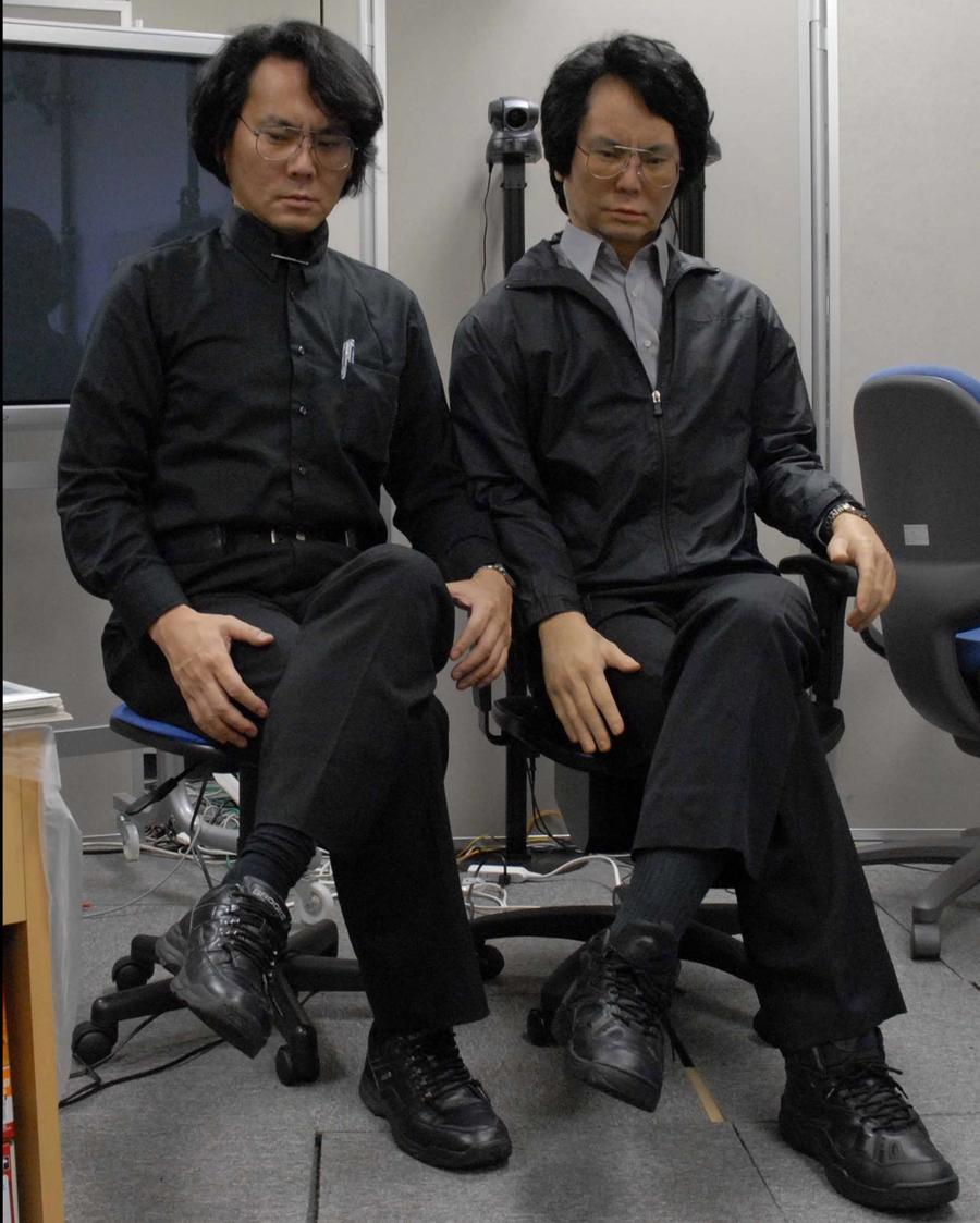 The roboticist and robot sit side by side in chairs with their legs crossed, arms on lap, and heads facing down.