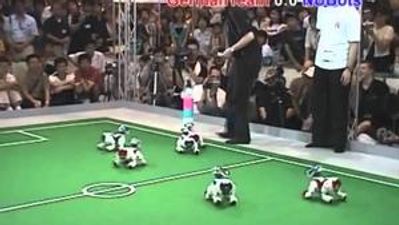 Five Aibo robot dogs play soccer on a small soccer field during a competition while an audience watches.