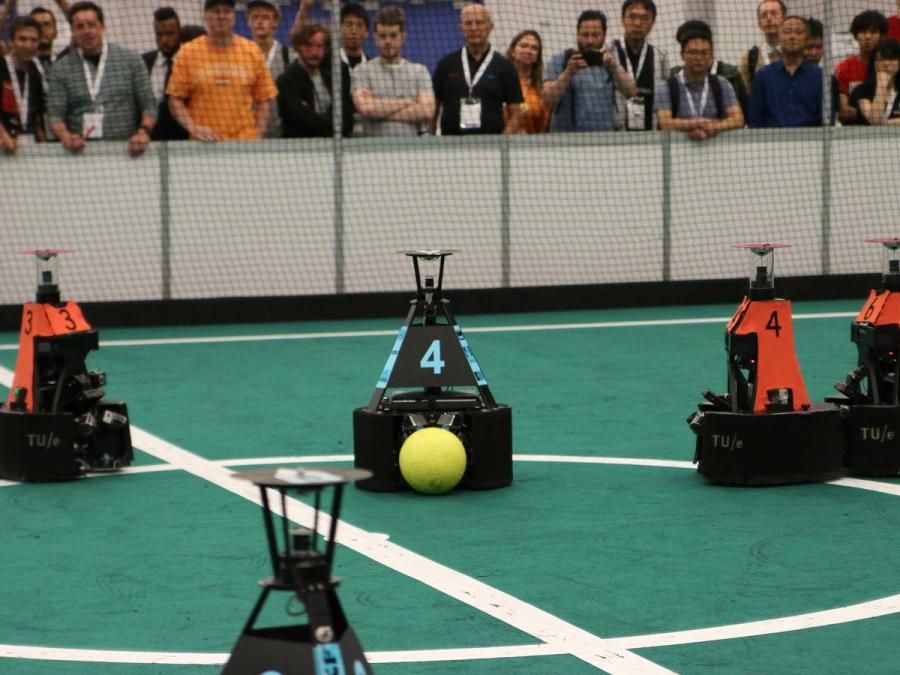 Two teams of mobile robots that consist of a triangular section on a black base play robots. One team wears orange and the other wears blue.