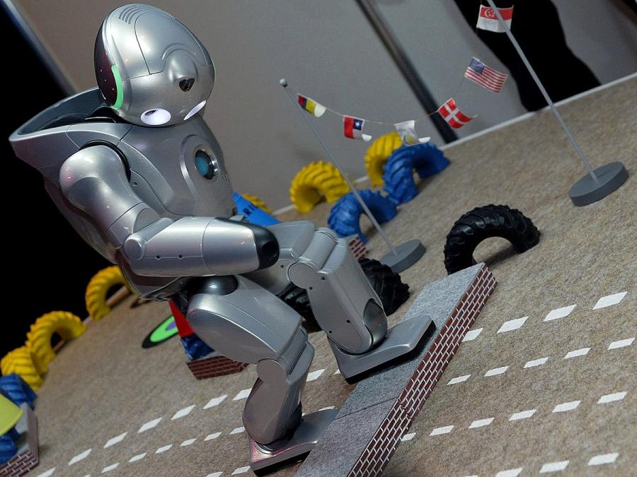 The silver humanoid robot looks down as it puts one foot up on a smal step.