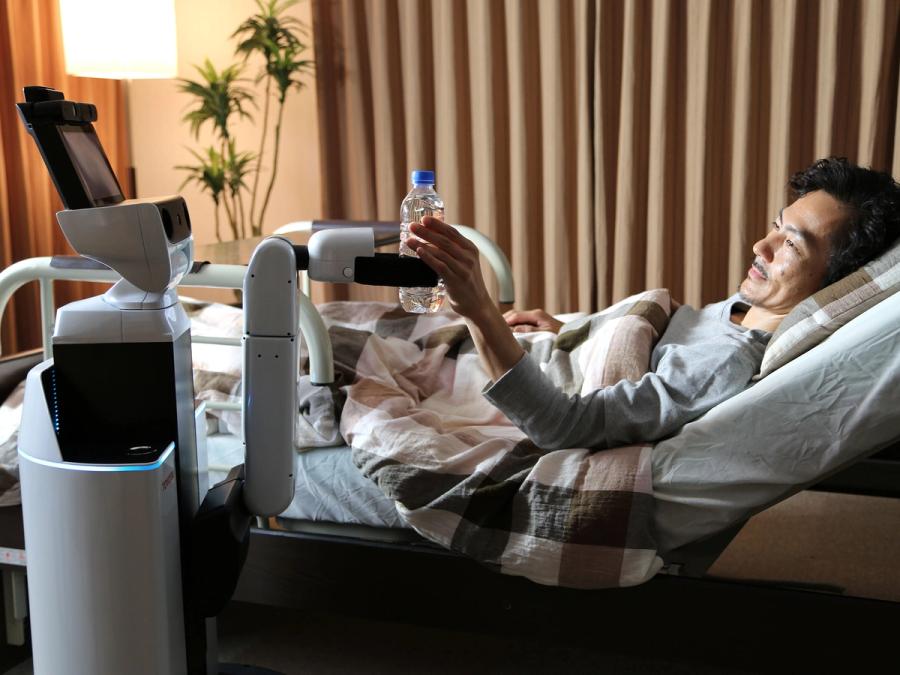 A mobile robot uses a claw hand on a single arm to hand a bottle of water to a patient in bed.