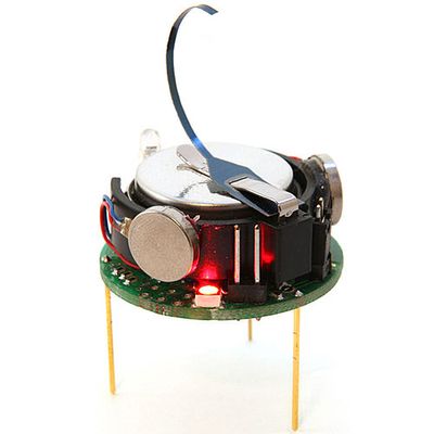 A single 5.5 cm tall kilobot which has 3 toothpick like yellow legs, electronics on a round circuit board and a curved metal piece sticking out of the top.