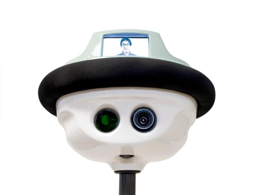Close up of QB's shiny white head with has two camera eyes, mouth, and the appearance of a hat, which has a screen showing a mans smiling face.