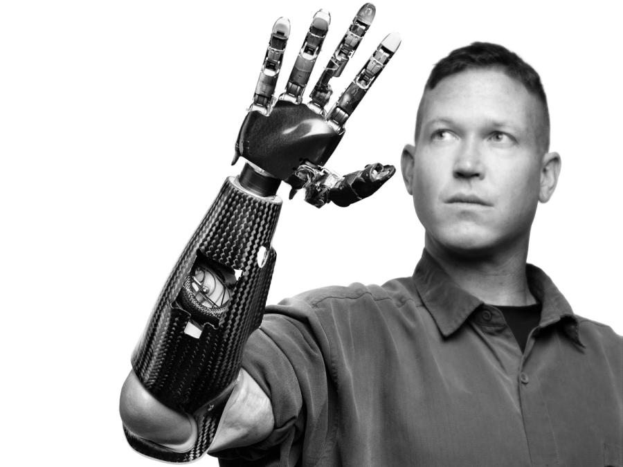 Black and white modern photograph of a man who is looking at his robotic arm and hand which he holds up in front of his face.