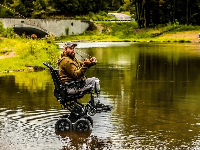 A man in a robotic wheelchair that is elevated on two of its four main wheels and sits in shallow water, holds a rod for fly fishing.