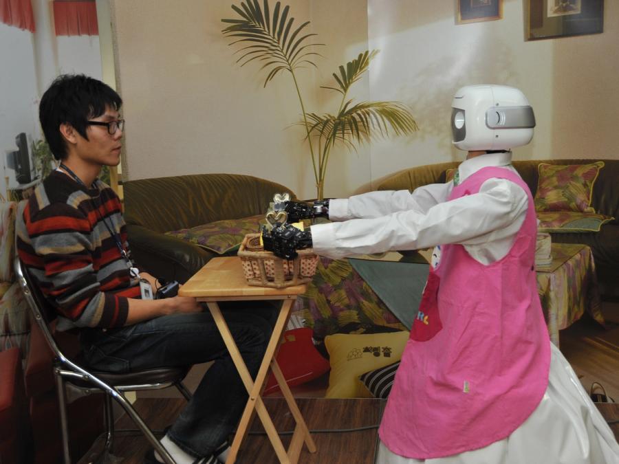 A humanoid robot wearing a pink apron serves a basket of food to a person sitting at a small table.