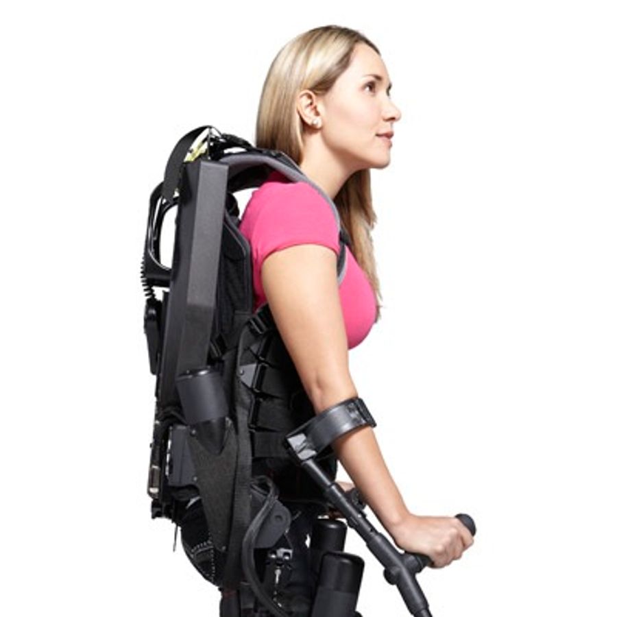 A blonde woman in a pink shirt wears a black exoskeleton and braces on her arms.