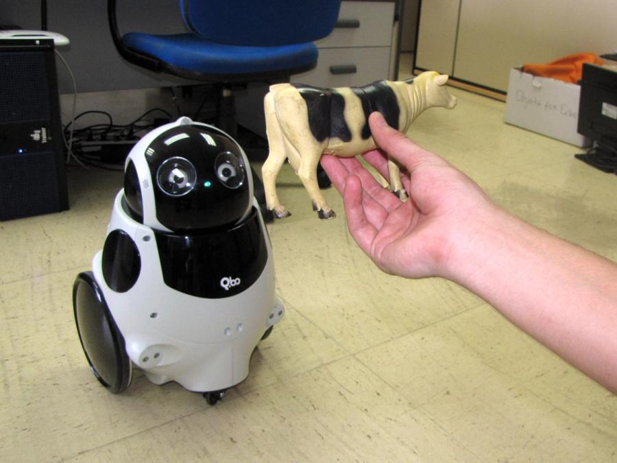 The robot looks up at a human hand, which is holding a toy cow.