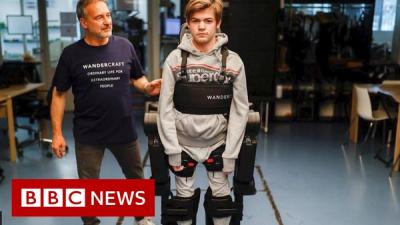 A young man wears the Atalante exoskeleton, which consists of a pair of robotic legs attached to the person's own legs, looking at the camera while another man stands nearby looking at him, with superimposed graphics showing the BBC News logo.