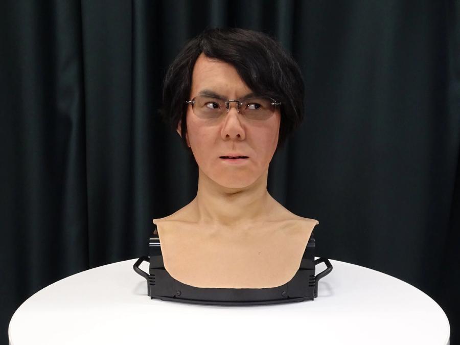 The robots head, with glasses, a mop of black hair, and an expressive face, is seen on a tabletop..