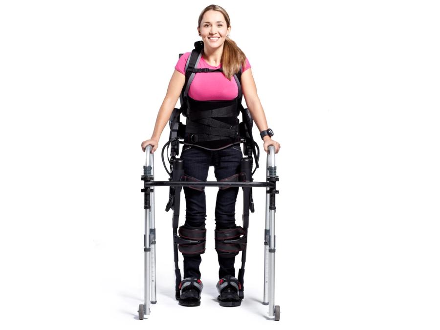 A smiling woman in a black exoskeleton suit holds onto a walker.