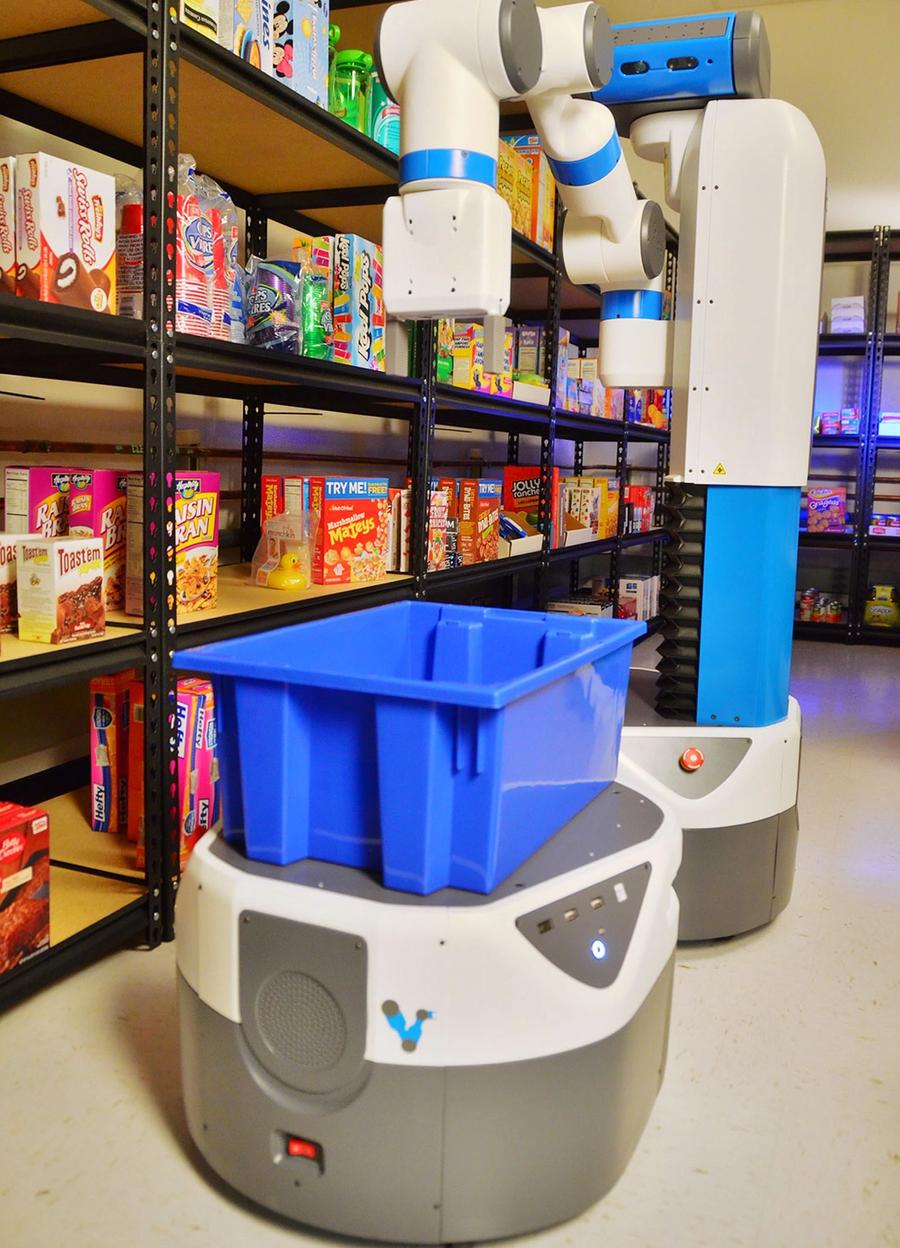 Two robots in front of a shelf of cereal in a store. A short mobile base carries a blue container while a tall mobile manipulation humanoid drops something into the container.