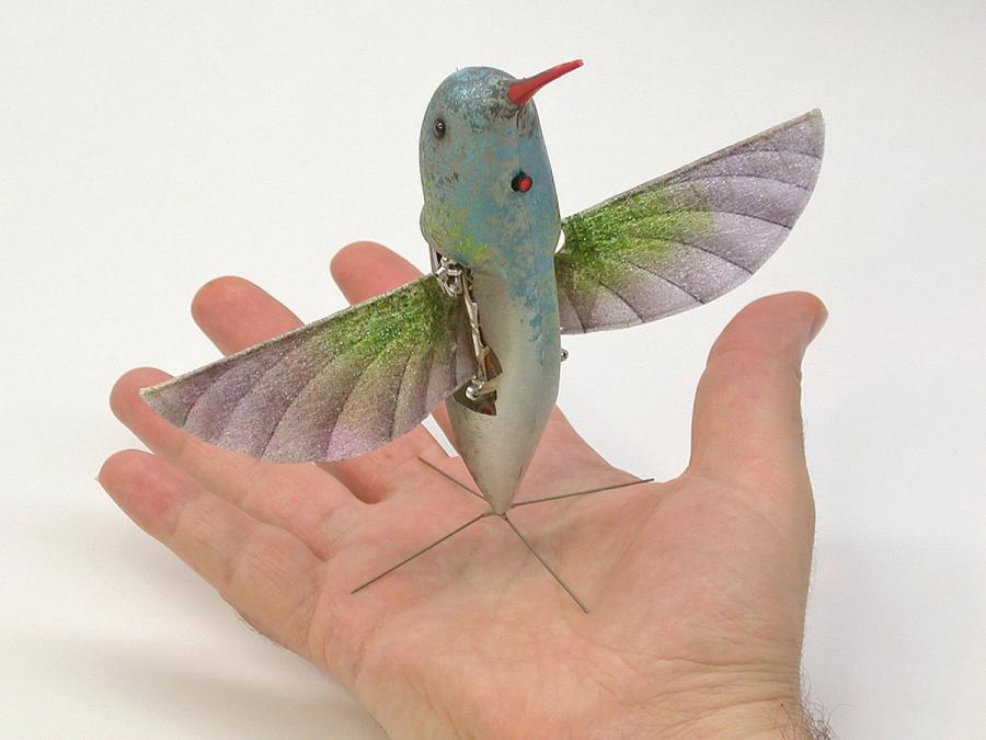 A robot that looks like a hummingbird including wings and long, thin, orange bill stands on a four pronged base on a human hand.