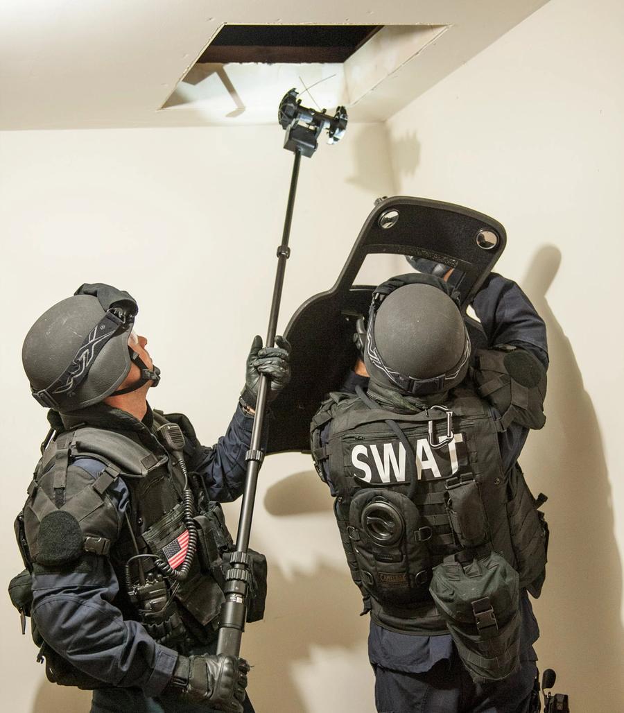 Two SWAT team members hold a pole with a Throwbot on the end up to a hole in a roof.