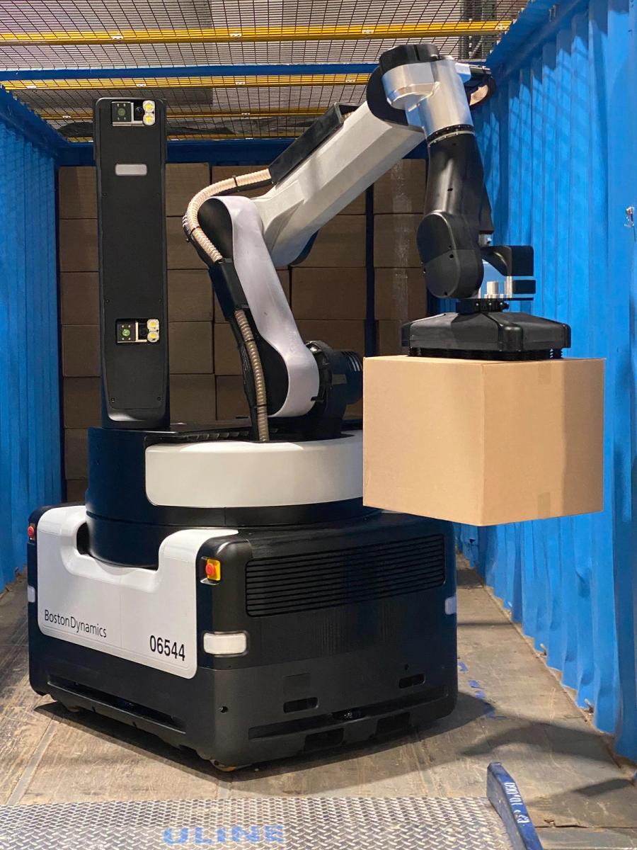 The robot sits in a blue shipping container, holding a cardboard box by the vacuum gripper at the end of its arm.