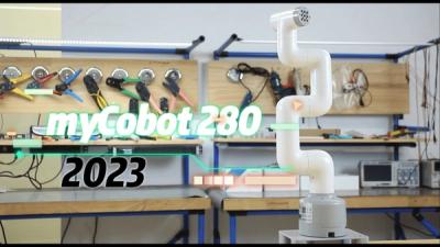 myCobot 280, a robot arm with a white plastic shell mounted on a metal base, stretches its links upwards in a lab with work benches and electronics equipment.