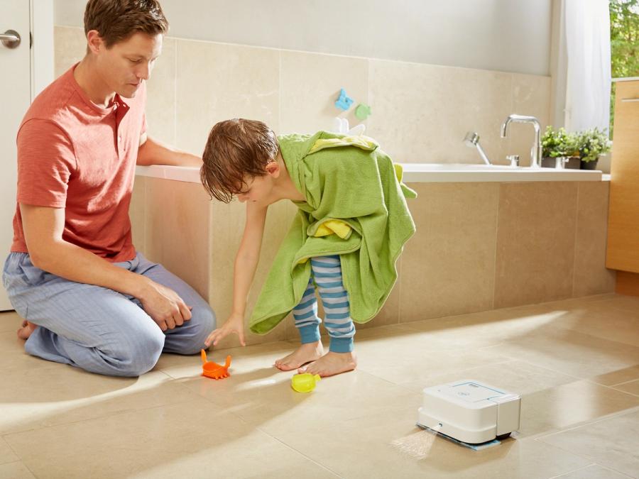 A small white robot mops around a father and child in a bathroom.