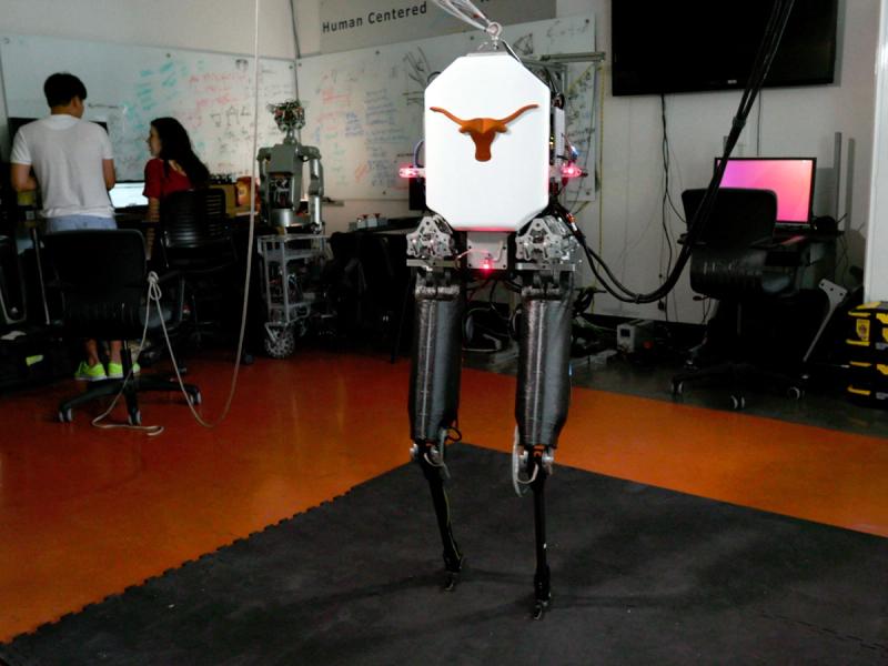 A bipedal robot with black legs and a white torso emblazoned with a longhorn silhouette stands in a lab.