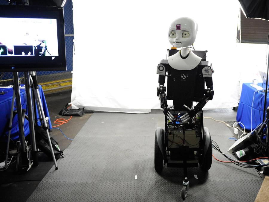 A black wheeled base and boxy torso support the robots expressive white face with eyes, nose, mouth, and a glowing square in its forehead. The robot has two arms with jointed human like silver fingers.