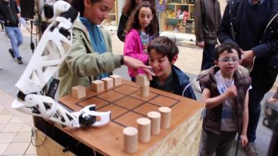 A plastic white robotic arm is mounted next to a tic-tac-toe board on the streets of Bordeaux, France, and a group of kids place pieces on the board to play the game.
