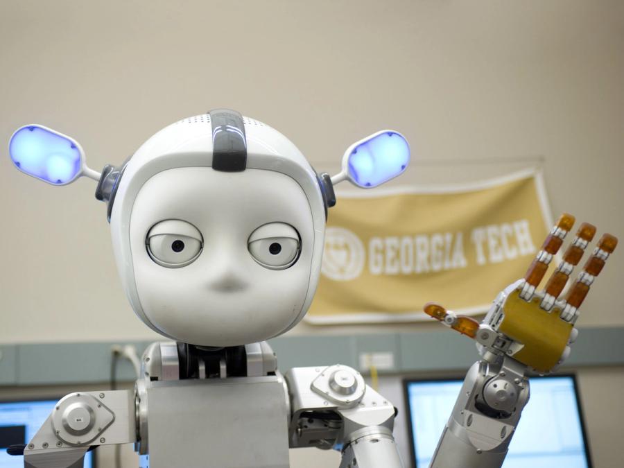 A robot with a round white face, two big eyes, and a helmet-like head shape with two large protruding ears that glow blue waves it's robotic four fingered hand.