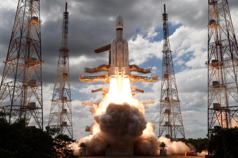 Chandrayaan-3 spacecraft launches, with a glowing yellow blast propelling it into a partly cloudy sky, surrounded by metal antenna towers.