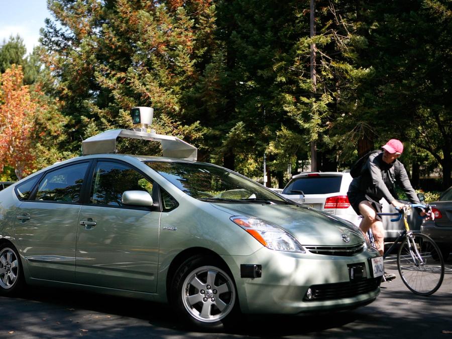A person riding a bike turns to look at a self driving car mounted with lidar and other equipment.