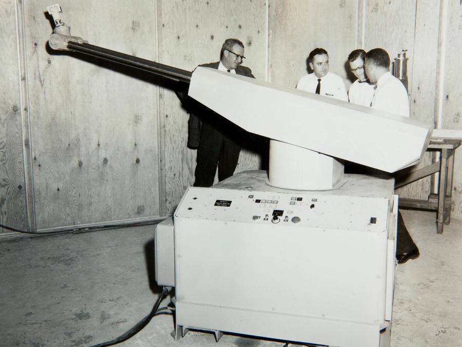 Black and white photo of a large robotic arm on a massive base that is almost as tall as the 4 men standing behind it.