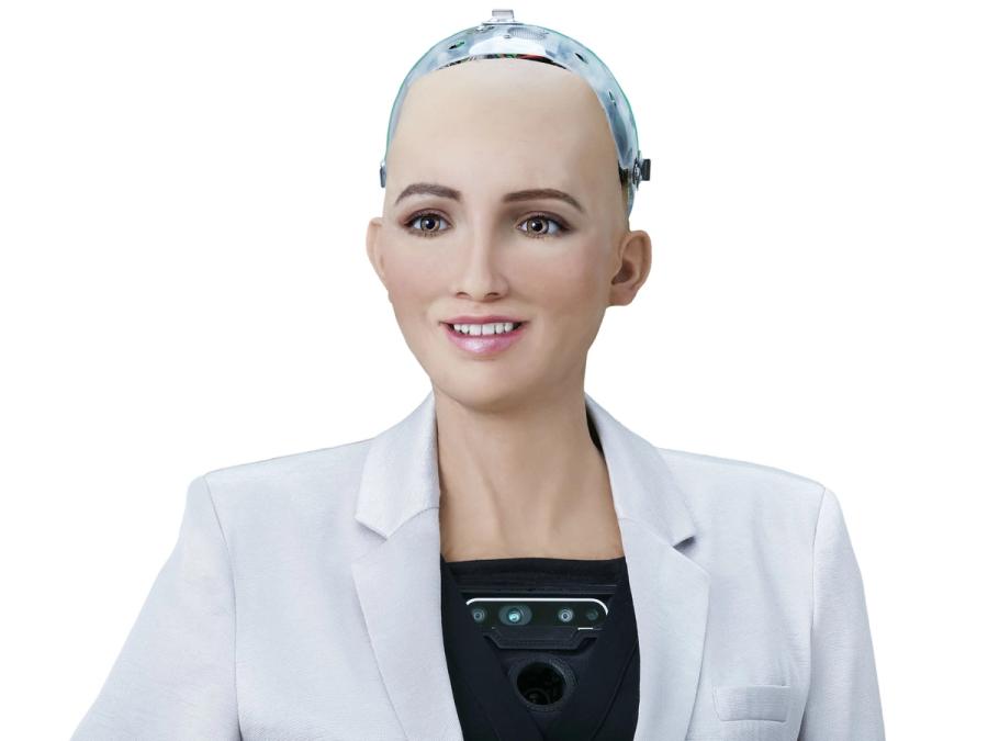 Sophia, a female appearing smiling robot with realistic peachy skin, green eyes, delicate nose, and pink lips with a tooth showing smile. The top of the robots head is a translucent cover housing electronics, and the robot wears a white blazer over her robotic torso, which includes a black plate with cameras and sensors.