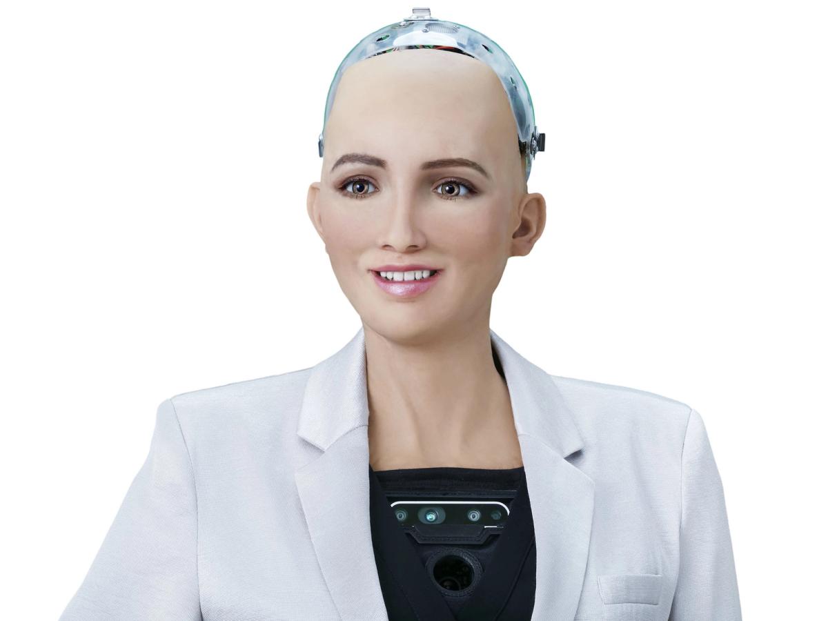 A female appearing smiling robot with realistic peachy skin, green eyes, delicate nose, and pink lips with a tooth showing smile. The top of the robots head is a translucent cover housing electronics, and the robot wears a white blazer over her robotic torso, which includes a black plate with cameras and sensors.