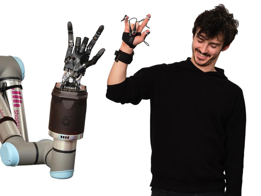 A robotic five fingered hand attached to a base gestures. To it's right is a smiling man doing the same gesture with his hands, wearing a special hand brace and sensors.
