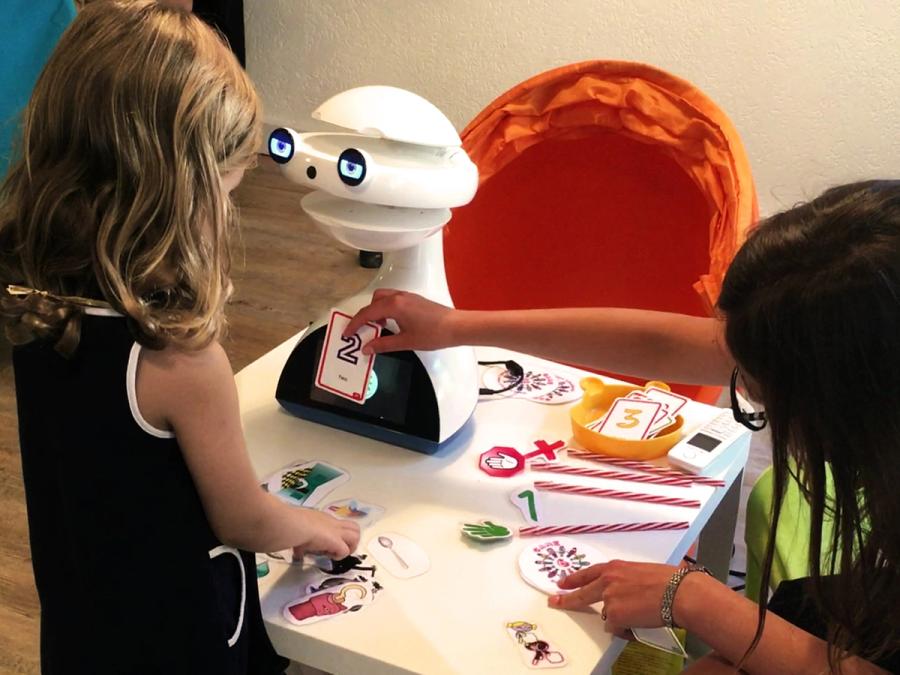 The robotic head sits on a table while two children interact with it by placing cards against its base.