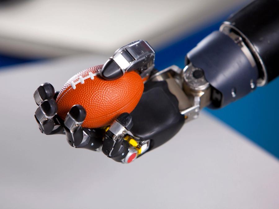 A black robotic hand with five jointed fingers mimicking a human hand holds an orange miniature football.