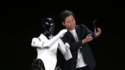A humanoid robot with a white and black plastic body stands next to a man who's taking a selfie of them both against a dark background.