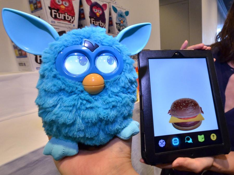 A blue furry Furby toy that resembles an owl, with large eyes, a yellow beak and pointy ears faces the camera, as does a tablet with an image of a cheeseburger.