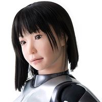 Close-up of a female robot with a realistic appearance including peach skin, pink lips, brown eyes and a black bob hairdo.