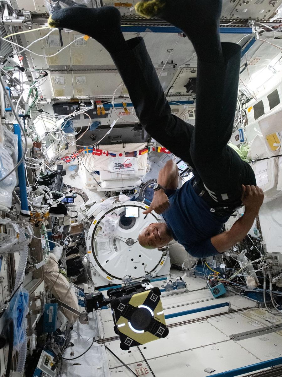 A smiling upside down astronaut floats with an Astrobee robot in a space station full of electronics.