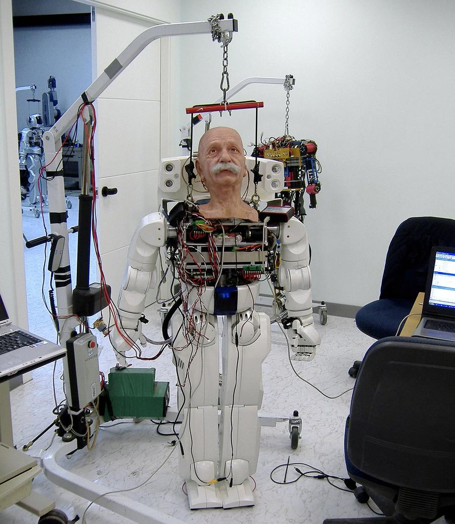 A male humanoid robot with a realistic face and white body held up by an overhead harness.