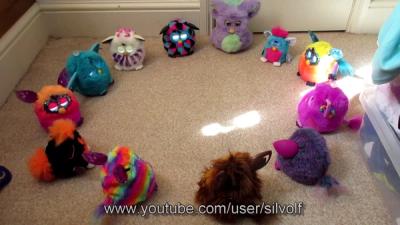 A dozen different multi color Furbys sit on a carpeted floor in a circle.