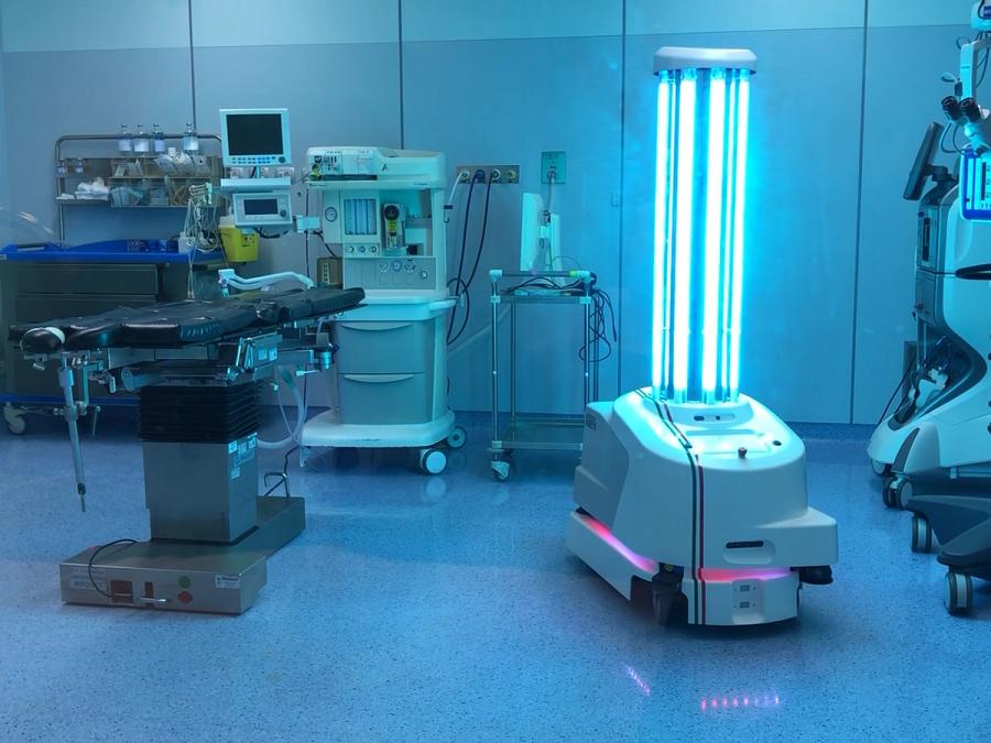 A wheeled mobile base with colorful lights inside supports a vertical column of brightly lit UV lightbulbs, inside of a surgical operation room.